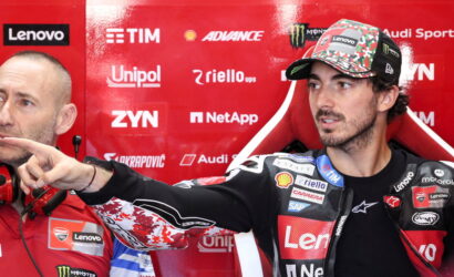 MotoGP rider in a Lenovo-branded racing suit and camo cap points to the left as a team member in red sits beside him in a sponsor-filled red backdrop.