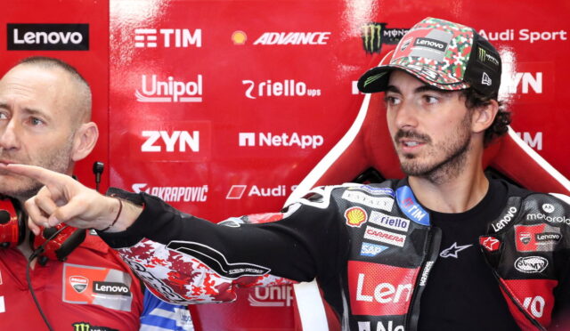 MotoGP rider in a Lenovo-branded racing suit and camo cap points to the left as a team member in red sits beside him in a sponsor-filled red backdrop.