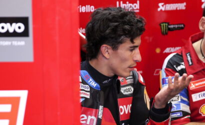 Side profile of a male motorcyclist in red/black racing gear in a paddock, sponsor banners in the background.