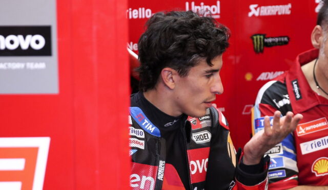 Side profile of a male motorcyclist in red/black racing gear in a paddock, sponsor banners in the background.
