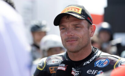 Male motorcycle racer wearing a Pirelli cap and sponsor-filled racing suit, speaking into a microphone during an interview in a crowded paddock.