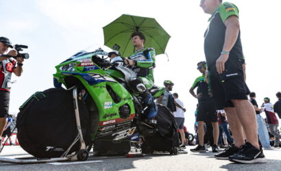 Rider in green Kawasaki racing leathers sits on a green motorcycle under a large green umbrella, surrounded by crew and photographers.