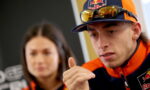 Man in Red Bull cap and orange-blue racing jacket gestures with his hand during a discussion, sunglasses resting on cap brim.