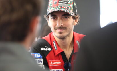 Man in a red sponsor-filled racing shirt being interviewed, wearing a colorful Lenovo cap and sunglasses hanging from his chest.