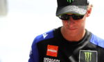 Male racer wearing a black cap with a neon-green Monster logo and dark sunglasses, looking downward.