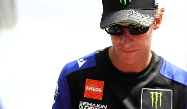 Male racer wearing a black cap with a neon-green Monster logo and dark sunglasses, looking downward.