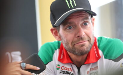 Man in a black Monster cap and green-red racing jersey gazes upward thoughtfully while holding a smartphone with a beaded bracelet visible on the wrist nearby.