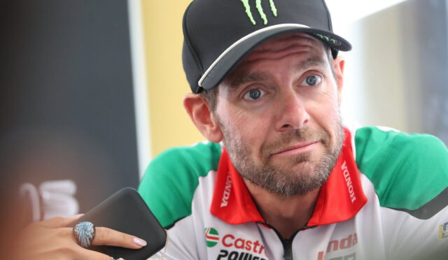 Man in a black Monster cap and green-red racing jersey gazes upward thoughtfully while holding a smartphone with a beaded bracelet visible on the wrist nearby.