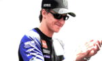 Motorcycle racer in blue Yamaha suit and cap, wearing sunglasses, applauding.