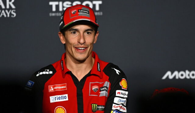 MotoGP rider in a red Ducati racing suit and cap, smiling at a media event.