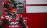 MotoGP rider in red Lenovo racing suit and helmet stands in the pit area with sponsor logos visible on gear and background signage.