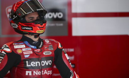 MotoGP rider in red Lenovo racing suit and helmet stands in the pit area with sponsor logos visible on gear and background signage.
