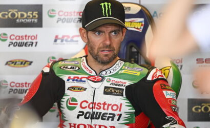 Racing rider in Castrol Power1 Honda suit sits in a pit area, wearing a black Monster cap and a focused expression amid sponsor banners behind him.” ,