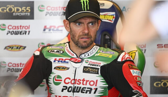 Racing rider in Castrol Power1 Honda suit sits in a pit area, wearing a black Monster cap and a focused expression amid sponsor banners behind him.” ,