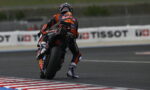Motorcycle racer in orange/black gear leans into a track turn on a racing bike with sponsor logos visible on suit and bike behind him.