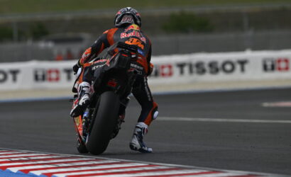 Motorcycle racer in orange/black gear leans into a track turn on a racing bike with sponsor logos visible on suit and bike behind him.