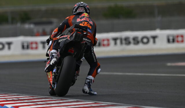 Motorcycle racer in orange/black gear leans into a track turn on a racing bike with sponsor logos visible on suit and bike behind him.