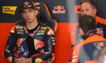 Moto rider in KTM/Red Bull racing suit sits in a black and orange racing chair, hands folded, in the pit area with team members nearby.