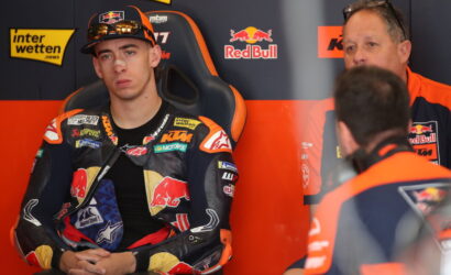 Moto rider in KTM/Red Bull racing suit sits in a black and orange racing chair, hands folded, in the pit area with team members nearby.