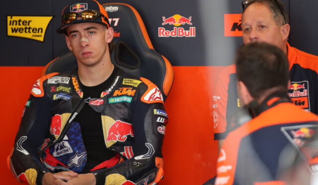 Moto rider in KTM/Red Bull racing suit sits in a black and orange racing chair, hands folded, in the pit area with team members nearby.