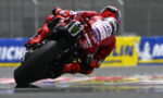 MotoGP rider in red gear leaning into a turn on a wet race track.