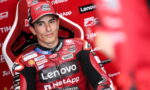 MotoGP rider in red Lenovo racing suit sitting in a red paddock chair, looking toward camera with sponsor logos on suit and backdrop