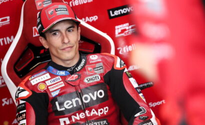 MotoGP rider in red Lenovo racing suit sitting in a red paddock chair, looking toward camera with sponsor logos on suit and backdrop