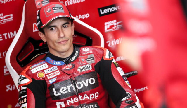 MotoGP rider in red Lenovo racing suit sitting in a red paddock chair, looking toward camera with sponsor logos on suit and backdrop