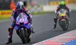 MotoGP rider on a blue Yamaha motorcycle leads a race on a curved track, with a rival in red pursuing behind.