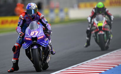 MotoGP rider on a blue Yamaha motorcycle leads a race on a curved track, with a rival in red pursuing behind.