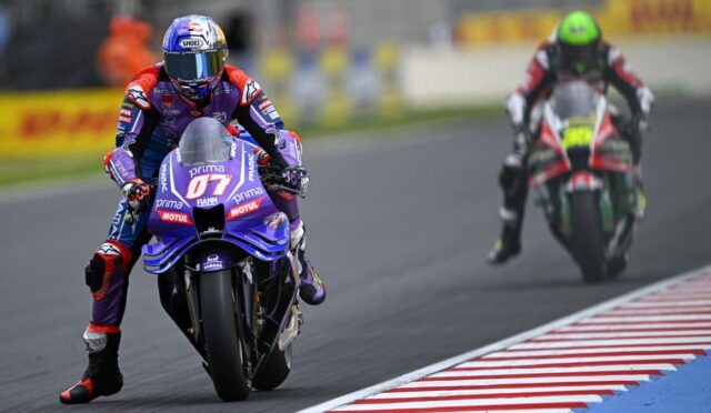 MotoGP rider on a blue Yamaha motorcycle leads a race on a curved track, with a rival in red pursuing behind.