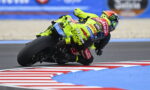 Motorbike racer in a neon-yellow suit leans into a sharp turn on a race track with red-and-white curbs nearby.