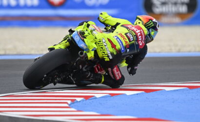 Motorbike racer in a neon-yellow suit leans into a sharp turn on a race track with red-and-white curbs nearby.