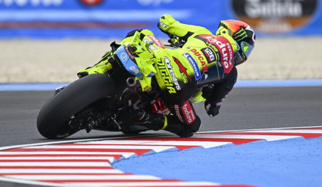 Motorbike racer in a neon-yellow suit leans into a sharp turn on a race track with red-and-white curbs nearby.