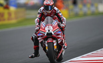 Motorcycle racer in red and white leathers riding a red race bike labeled number 63 on a track with sponsor stickers visible on the bike and suit.