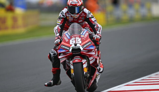 Motorcycle racer in red and white leathers riding a red race bike labeled number 63 on a track with sponsor stickers visible on the bike and suit.