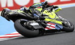 Two riders on sport bikes lean into a turn on a racetrack; neon yellow bike features flag decals and bold graphics.