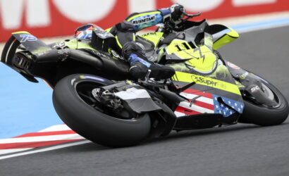 Two riders on sport bikes lean into a turn on a racetrack; neon yellow bike features flag decals and bold graphics.