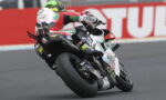 Motorcycle racer wearing red, white, and green leans into a turn on a sportbike during a race.