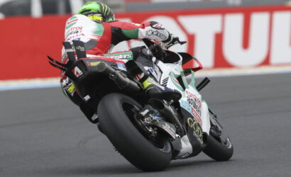 Motorcycle racer wearing red, white, and green leans into a turn on a sportbike during a race.
