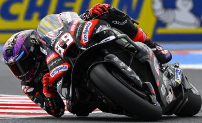 MotoGP rider leaning into a turn on a black-red racing bike, number 89, with sponsor logos visible on the suit and bike