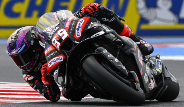 MotoGP rider leaning into a turn on a black-red racing bike, number 89, with sponsor logos visible on the suit and bike