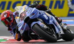 MotoGP rider on a blue racing bike leans hard into a left-hand turn, helmet red and track barriers in the background.