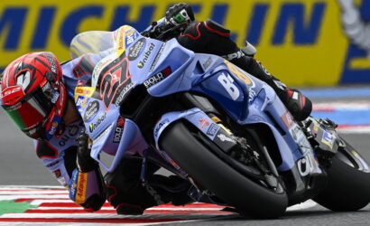 MotoGP rider on a blue racing bike leans hard into a left-hand turn, helmet red and track barriers in the background.