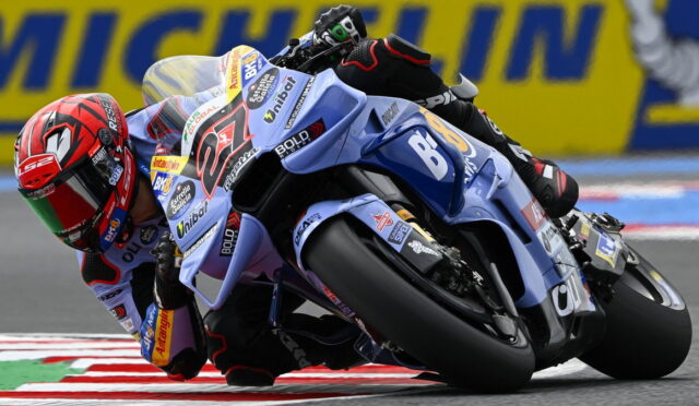 MotoGP rider on a blue racing bike leans hard into a left-hand turn, helmet red and track barriers in the background.
