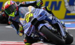 Motorcycle racer in a bright helmet leaning into a turn on a blue racing bike during a race (number 84).