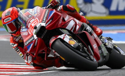 MotoGP rider in red suit and helmet leaning into a sharp corner on a red Ducati bike with sponsor decals (Lenovo, Shell) in a race track setting
