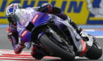 Motorcycle racer leaning into a tight turn on a purple race bike with sponsor logos, close to the curb.