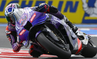 Motorcycle racer leaning into a tight turn on a purple race bike with sponsor logos, close to the curb.