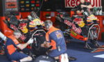 Two Red Bull KTM factory racing motorcycles are on stands in a busy pit garage as crew members service the bikes and check tires behind them.